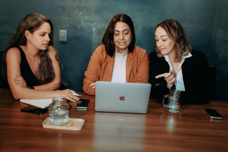 Three women working together on a laptop, discussing a project in a professional setting.